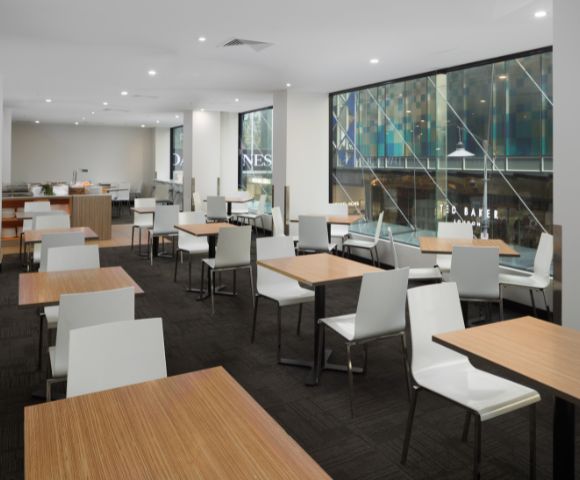 Bright, modern café with light wood tables and white chairs. Large windows reveal an urban street view, creating an open and inviting atmosphere.