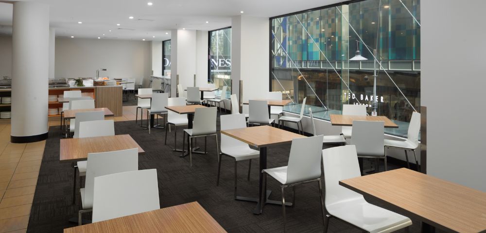 Bright, modern café with light wood tables and white chairs. Large windows reveal an urban street view, creating an open and inviting atmosphere.