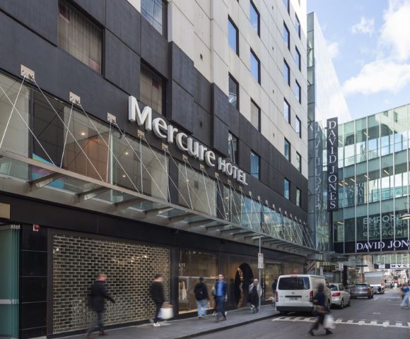 Street view of Mercure Hotel's modern facade on a bustling city street. Pedestrians walk by, and parked cars line the road. Storefronts are visible.