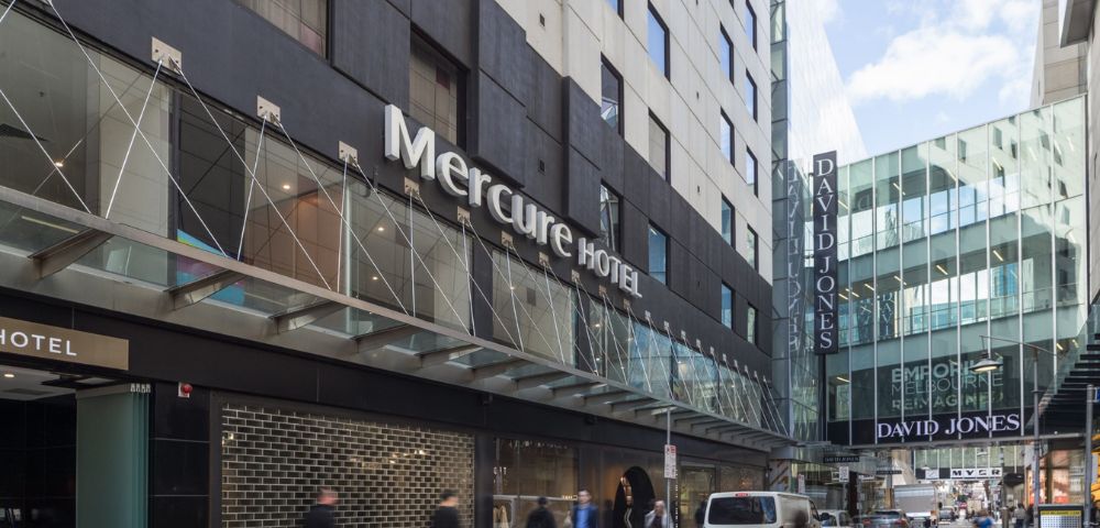 Street view of Mercure Hotel's modern facade on a bustling city street. Pedestrians walk by, and parked cars line the road. Storefronts are visible.