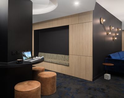 Modern lounge area with black walls and soft lighting. Features a cozy alcove with a bench, round leather stools, and a laptop on a curved table.