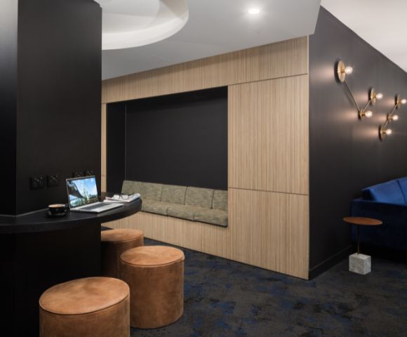 Modern lounge area with black walls and soft lighting. Features a cozy alcove with a bench, round leather stools, and a laptop on a curved table.