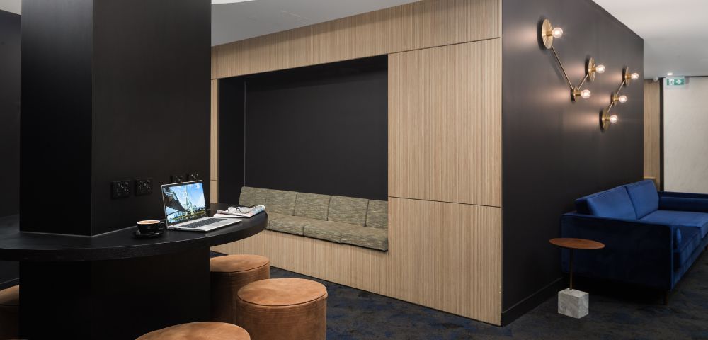 Modern lounge area with black walls and soft lighting. Features a cozy alcove with a bench, round leather stools, and a laptop on a curved table.