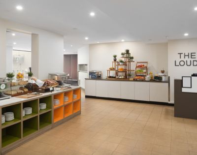 Bright, modern breakfast buffet area with a variety of pastries, fruits, and plates on display. Shelving is organized and inviting, creating a welcoming atmosphere.
