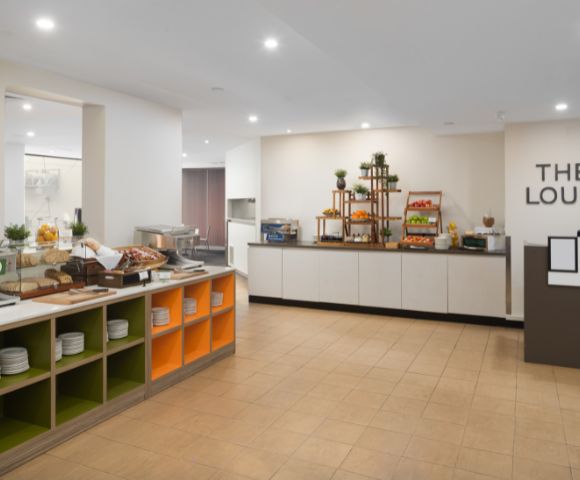 Bright, modern breakfast buffet area with a variety of pastries, fruits, and plates on display. Shelving is organized and inviting, creating a welcoming atmosphere.