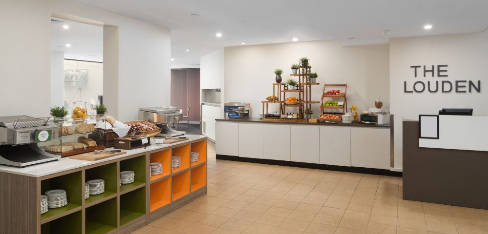 Bright, modern breakfast buffet area with a variety of pastries, fruits, and plates on display. Shelving is organized and inviting, creating a welcoming atmosphere.