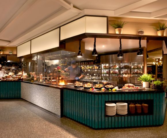 Modern buffet restaurant interior with teal counters, hanging lamps, and a diverse food spread. Chefs work behind glass panels, creating a lively ambiance.