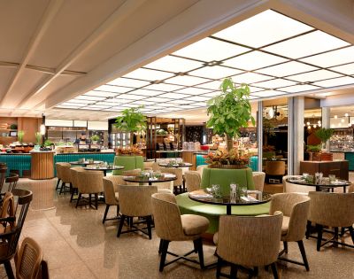 A modern restaurant with round tables and beige chairs, lush plants as decor, and a softly lit buffet area, creating a warm, inviting atmosphere.
