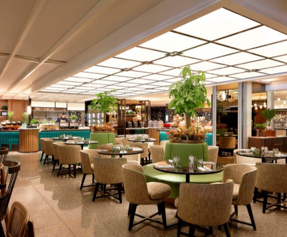 A modern restaurant with round tables and beige chairs, lush plants as decor, and a softly lit buffet area, creating a warm, inviting atmosphere.