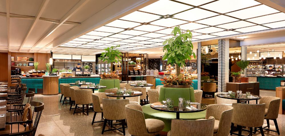 A modern restaurant with round tables and beige chairs, lush plants as decor, and a softly lit buffet area, creating a warm, inviting atmosphere.