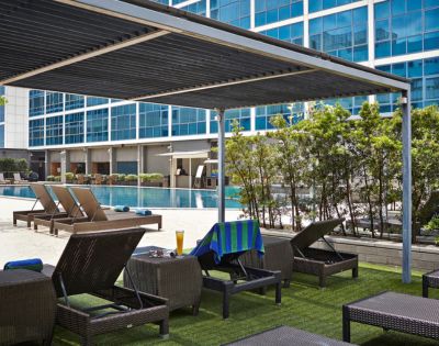 Poolside lounge area with wicker chairs and a blue towel, under a sleek pergola. Nearby, a pool reflects a modern glass building facade.