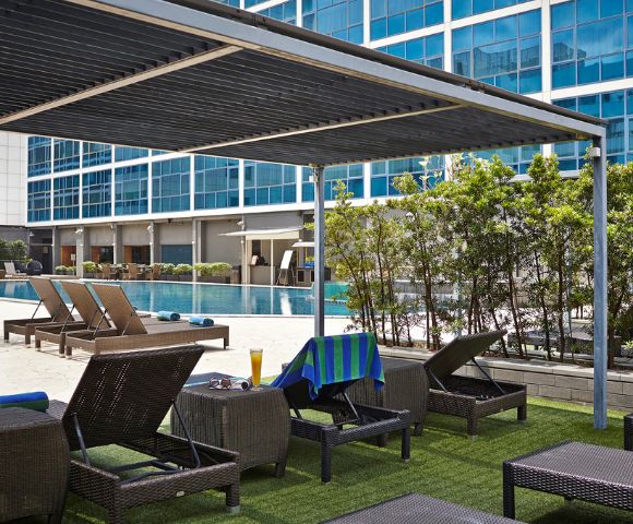 Poolside lounge area with wicker chairs and a blue towel, under a sleek pergola. Nearby, a pool reflects a modern glass building facade.