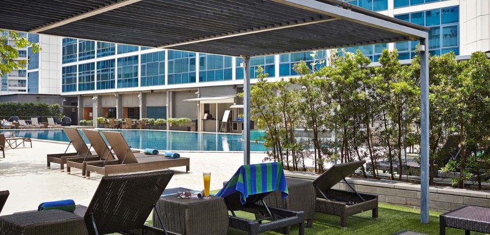 Poolside lounge area with wicker chairs and a blue towel, under a sleek pergola. Nearby, a pool reflects a modern glass building facade.