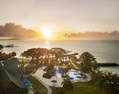 Sunset view of a coastal resort with a pool, surrounded by palm trees. Calm ocean in the background and vibrant sky, creating a serene atmosphere.