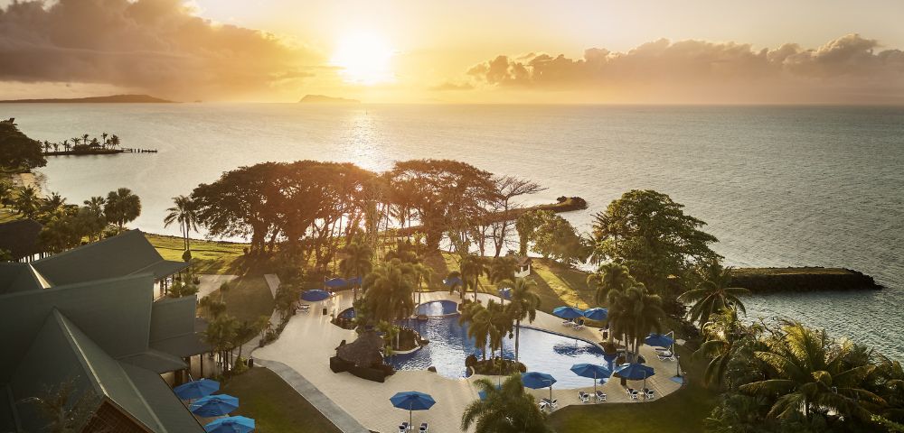 Sunset view of a coastal resort with a pool, surrounded by palm trees. Calm ocean in the background and vibrant sky, creating a serene atmosphere.