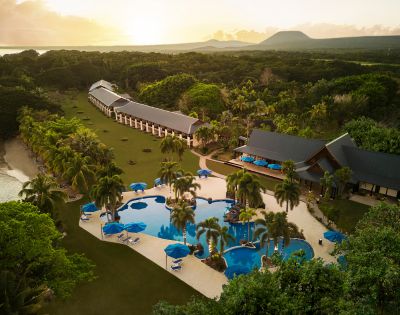 Aerial view of a tropical resort at sunset, featuring a curvy pool with palm trees, sun loungers, and oceanfront villas surrounded by lush greenery.