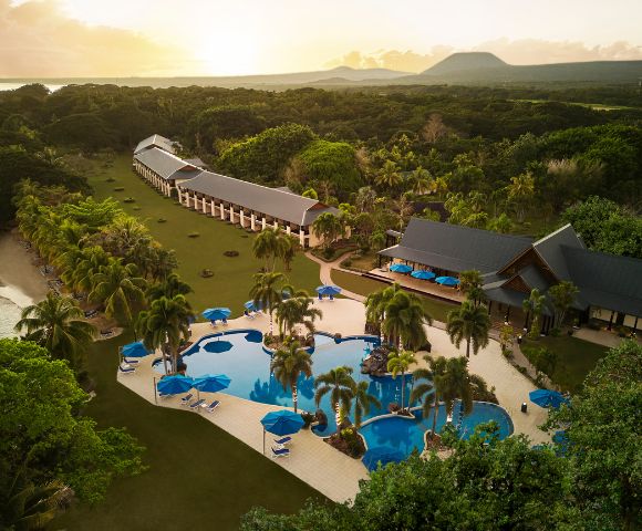 Aerial view of a tropical resort at sunset, featuring a curvy pool with palm trees, sun loungers, and oceanfront villas surrounded by lush greenery.