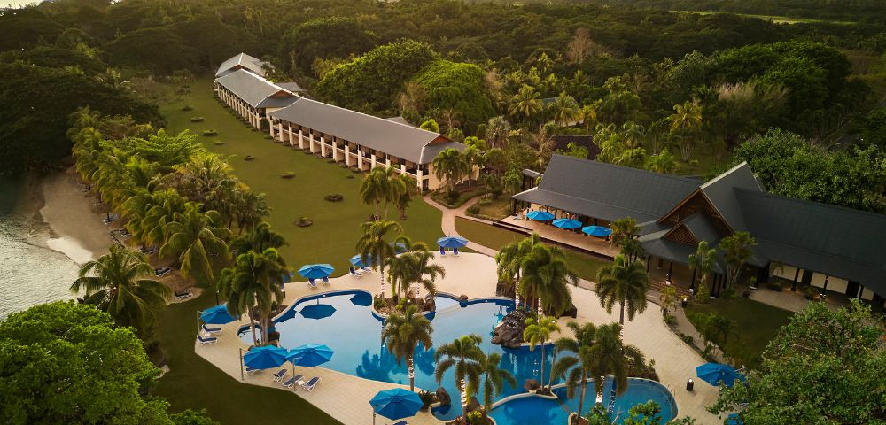 Aerial view of a tropical resort at sunset, featuring a curvy pool with palm trees, sun loungers, and oceanfront villas surrounded by lush greenery.