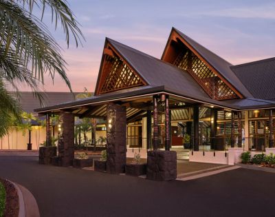 A modern hotel entrance at dusk with angled roofs and warm lighting. Palm trees frame the scene, adding a tropical feel. The sky is softly lit with pink hues.