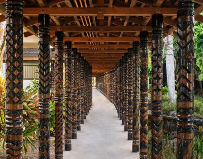 A covered walkway with intricately carved wooden pillars, featuring geometric patterns. Sunlight filters through, creating a serene, inviting atmosphere.