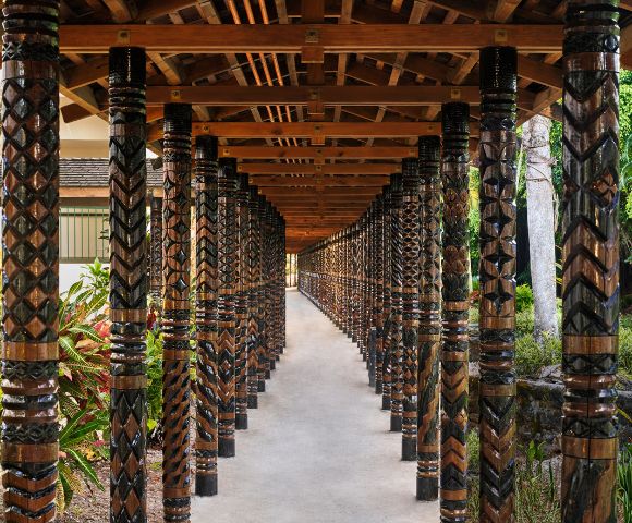 A covered walkway with intricately carved wooden pillars, featuring geometric patterns. Sunlight filters through, creating a serene, inviting atmosphere.