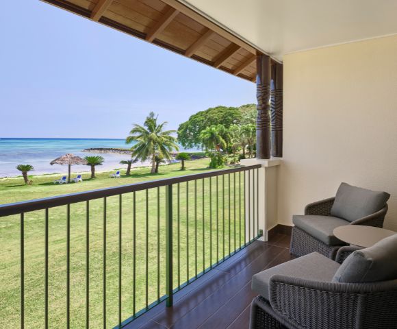 Balcony with two wicker chairs and a small table overlooking a grassy lawn with palm trees, thatched umbrellas, and a tranquil ocean under a clear sky.