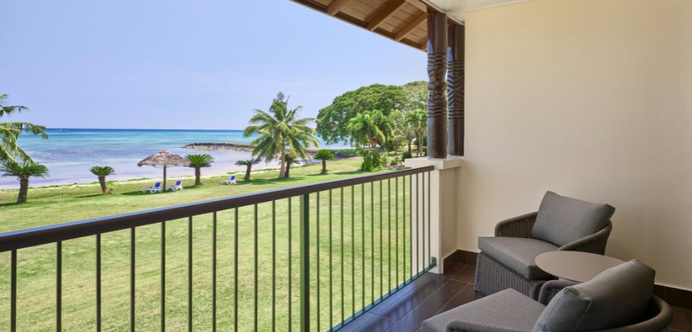 Balcony with two wicker chairs and a small table overlooking a grassy lawn with palm trees, thatched umbrellas, and a tranquil ocean under a clear sky.