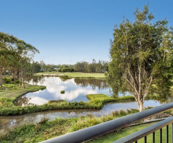 Tranquil scene of a pond surrounded by lush greenery and trees, reflecting a clear blue sky, viewed from a balcony. Peaceful and serene atmosphere.