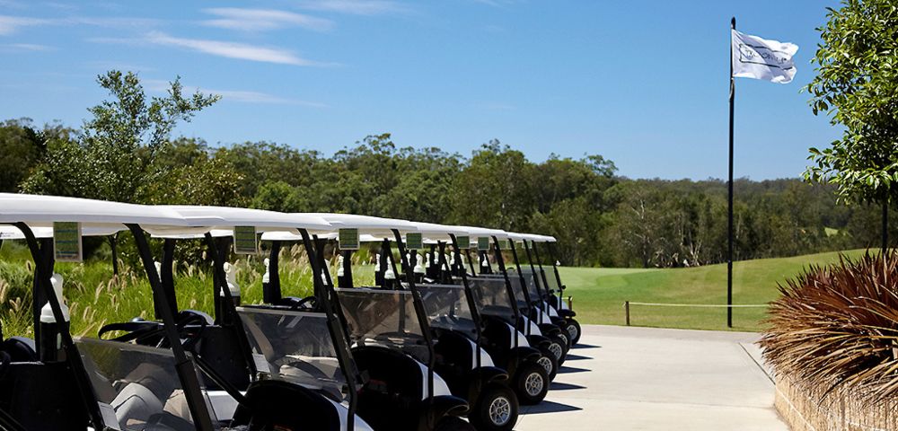 A row of golf carts parked on a sunlit path by a lush green golf course, with a flag waving on a pole and clear blue sky above.