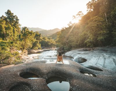 A person sits on rocky terrain with circular water pools, facing a sunlit forest and mountains. The scene is serene and bathed in warm sunlight.