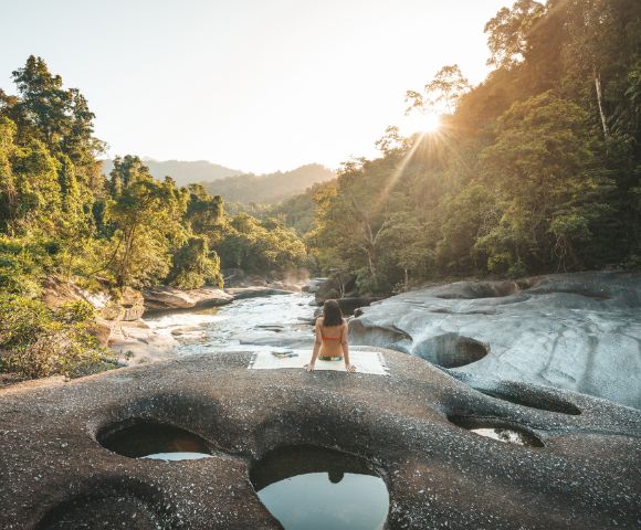 A person sits on rocky terrain with circular water pools, facing a sunlit forest and mountains. The scene is serene and bathed in warm sunlight.