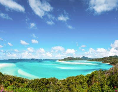 Vibrant tropical beach with turquoise waters, lush greenery, and a bright blue sky with scattered clouds. The scene conveys tranquility and natural beauty.