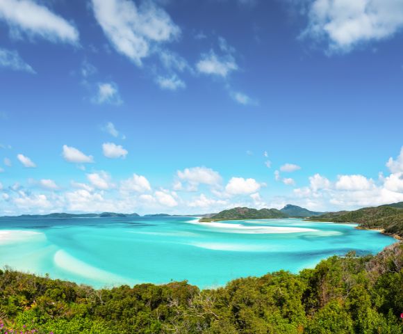 Vibrant tropical beach with turquoise waters, lush greenery, and a bright blue sky with scattered clouds. The scene conveys tranquility and natural beauty.