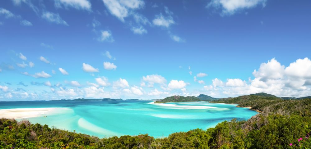 Vibrant tropical beach with turquoise waters, lush greenery, and a bright blue sky with scattered clouds. The scene conveys tranquility and natural beauty.