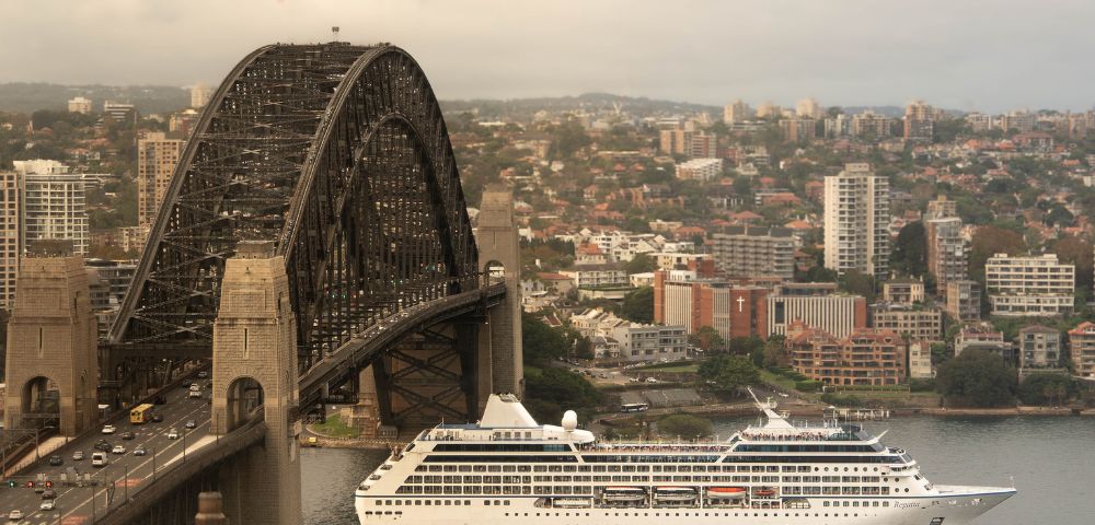A cruise ship sails under a large steel bridge in an urban area. The cityscape background has dense buildings and a slightly overcast sky, evoking calmness.