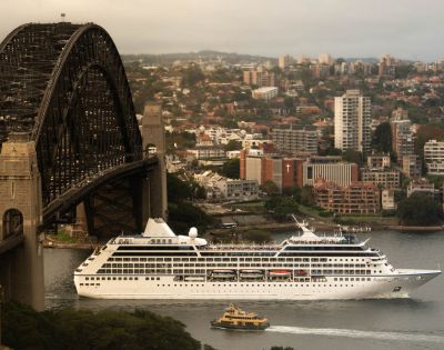 A cruise ship sails under a large steel bridge in an urban area. The cityscape background has dense buildings and a slightly overcast sky, evoking calmness.