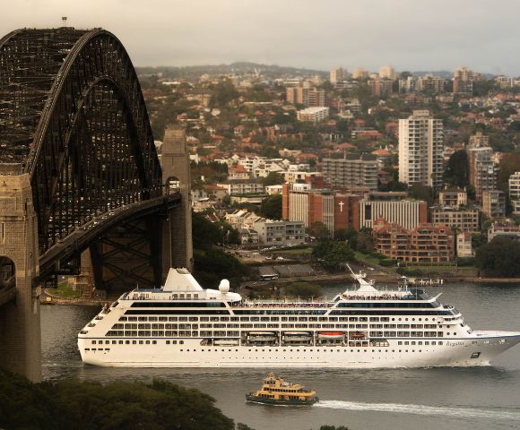 A cruise ship sails under a large steel bridge in an urban area. The cityscape background has dense buildings and a slightly overcast sky, evoking calmness.
