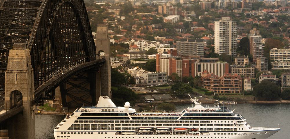 A cruise ship sails under a large steel bridge in an urban area. The cityscape background has dense buildings and a slightly overcast sky, evoking calmness.