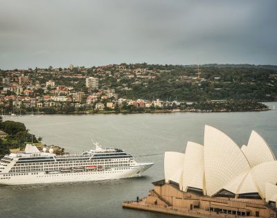 A white cruise ship sails past Sydney Opera House, with its iconic sails in the foreground. The cityscape and green hills form the backdrop. Calm, scenic view.