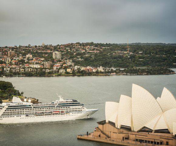 A white cruise ship sails past Sydney Opera House, with its iconic sails in the foreground. The cityscape and green hills form the backdrop. Calm, scenic view.