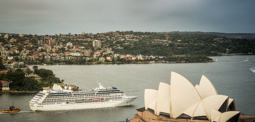 A white cruise ship sails past Sydney Opera House, with its iconic sails in the foreground. The cityscape and green hills form the backdrop. Calm, scenic view.