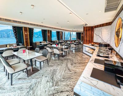 Bright, elegant restaurant with marble floors and tables, overlooking a city skyline through large windows. Sophisticated ambiance with a buffet counter.