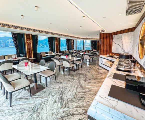 Bright, elegant restaurant with marble floors and tables, overlooking a city skyline through large windows. Sophisticated ambiance with a buffet counter.