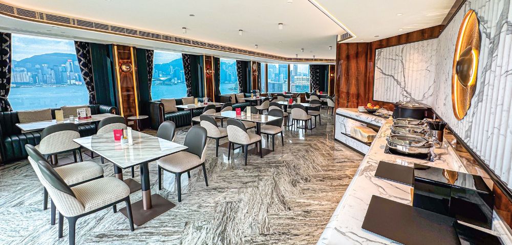 Bright, elegant restaurant with marble floors and tables, overlooking a city skyline through large windows. Sophisticated ambiance with a buffet counter.
