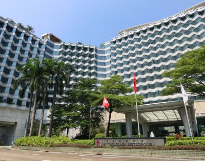 Modern high-rise hotel with a wave-like facade, surrounded by lush greenery and flags near the entrance. Clear blue sky adds a peaceful tone.
