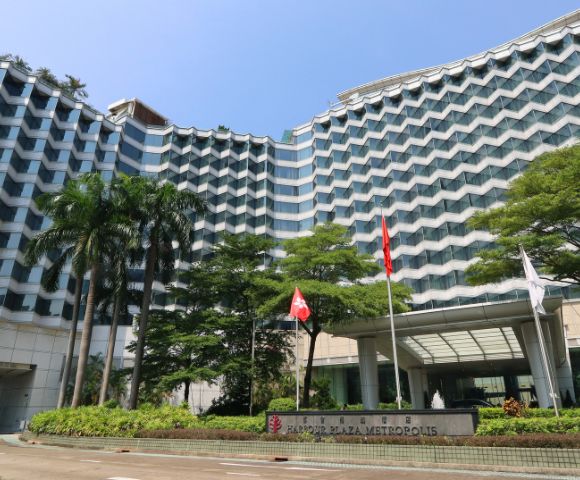 Modern high-rise hotel with a wave-like facade, surrounded by lush greenery and flags near the entrance. Clear blue sky adds a peaceful tone.