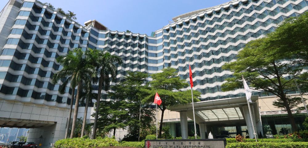 Modern high-rise hotel with a wave-like facade, surrounded by lush greenery and flags near the entrance. Clear blue sky adds a peaceful tone.