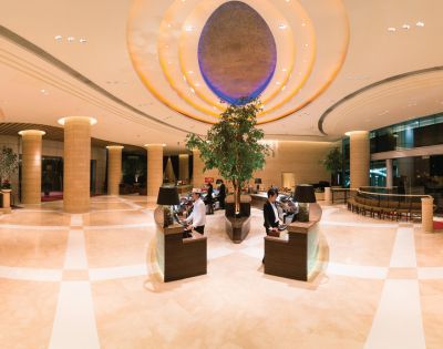 Luxurious hotel lobby with high ceilings, elegant lighting, and warm tones. Two people sit at sleek desks, surrounded by large plants and modern decor.
