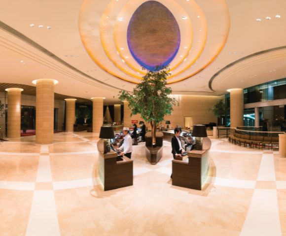 Luxurious hotel lobby with high ceilings, elegant lighting, and warm tones. Two people sit at sleek desks, surrounded by large plants and modern decor.