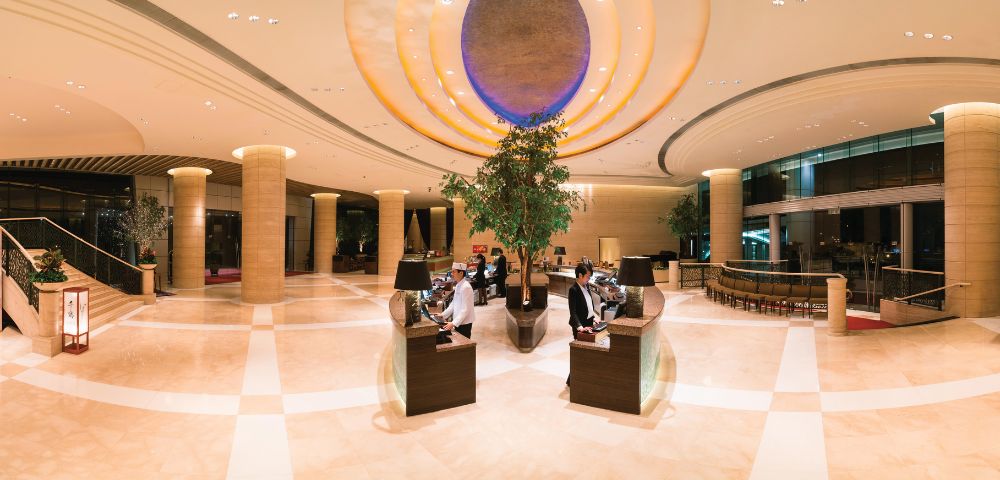 Luxurious hotel lobby with high ceilings, elegant lighting, and warm tones. Two people sit at sleek desks, surrounded by large plants and modern decor.